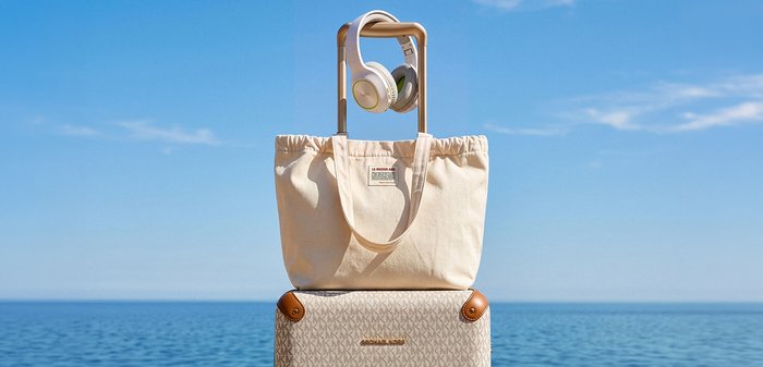Sac fourre-tout beige avec étiquette et casque suspendu à la poignée d'une valise de voyage au bord d'une mer bleue calme sous un ciel dégagé.