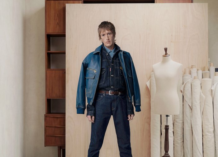 Άνδρας στέκεται με τζιν μπουφάν και τζιν παντελόνι δίπλα σε μανεκέν, με ρολά υφάσματος και ξύλινα ράφια σε ένα στούντιο.