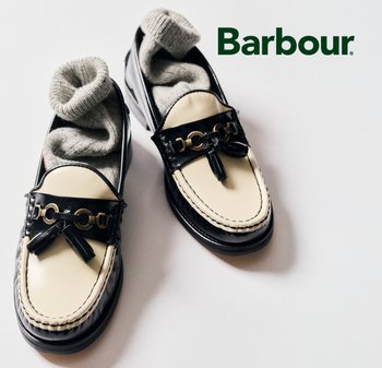 Schwarze und cremefarbene Loafers mit glänzender Oberfläche, die mit einer goldenen Kettenakzent und einer Schleifendetails versehen sind. Graue Socken sind teilweise sichtbar.