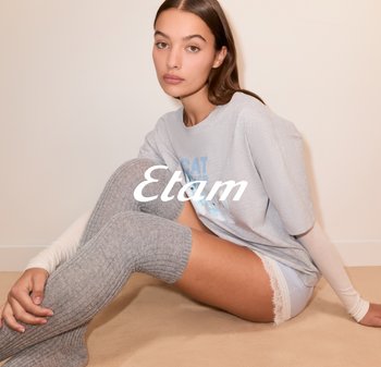 Graues übergroßes T-Shirt mit blauer Schrift, weiße Spitzenverzierung an den Shorts und knielangen, gerippten grauen Socken, auf einer hellbeigen Oberfläche.