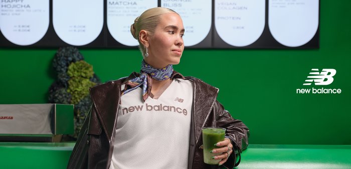 Vrouw met blond haar in een leren jas en sjaal houdt een groene drank vast, staand tegen een groene muur met het New Balance-logo.