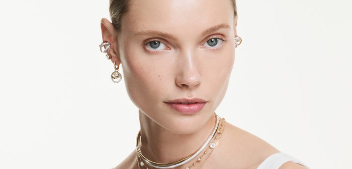 Vrouw met lichte huid en blauwe ogen draagt gelaagde gouden en diamanten oorbellen en kettingen, gekleed in een wit mouwloos topje, neutrale achtergrond.