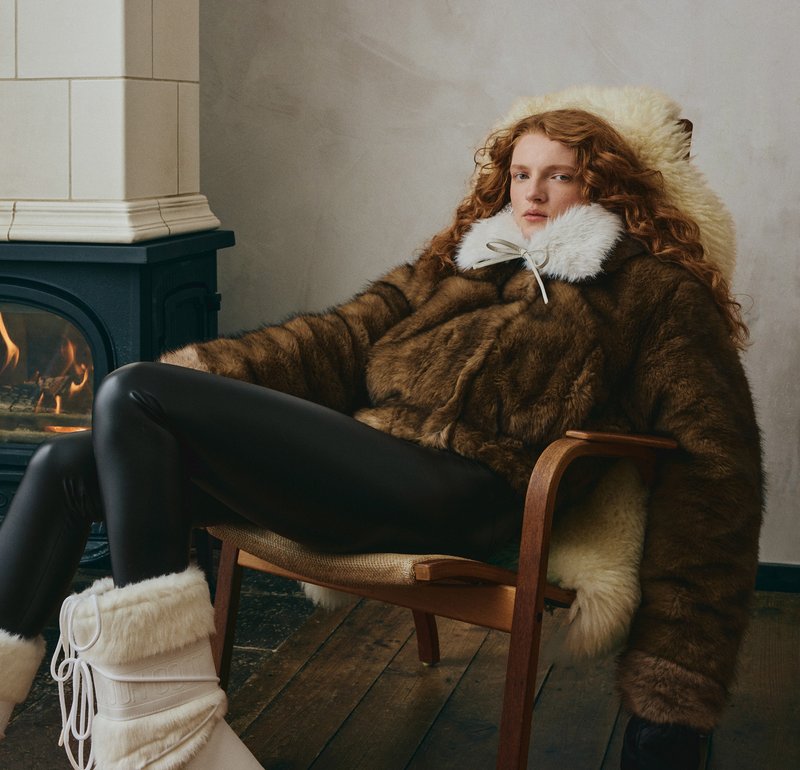 Schwarze übergroße Sonnenbrille, flauschiger weißer Kragen, brauner Pelzmantel und hängende Kristallohrringe erzeugen einen markanten, strukturierten Look.