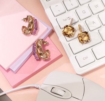 Ohrringe aus Gold und lila Edelsteinen mit kantigen Formen und klaren Akzenten, auf pastellfarbenem Papier und einer weißen Tastatur neben einer Maus platziert.