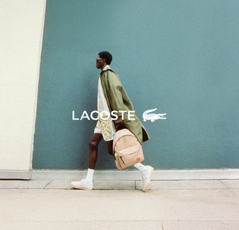 Plecak marki Lacoste o miękkim beżowym wykończeniu. Model ma na sobie zielony płaszcz na lekkich wzorzystych szortach, białych skarpetkach i trampkach.