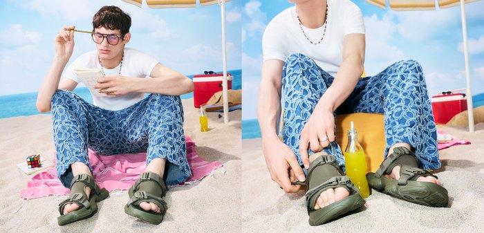 Jong persoon zit op een strandlaken onder een parasol, draagt een broek met patroon en sandalen, houdt een potlood en notitieboekje vast.
