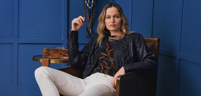 Vrouw met golvend blond haar zit zelfverzekerd in een leren stoel, gekleed in een zwart doorgestikt jack, een top met kettingpatroon en witte broek, tegen een blauwe achtergrond met panelen.