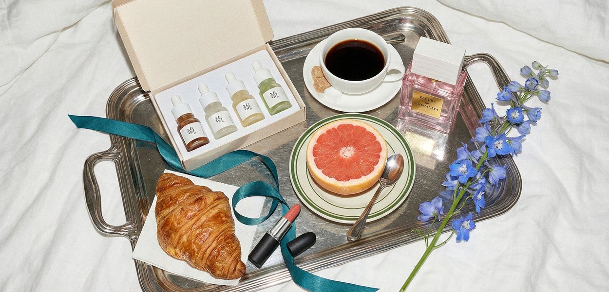 Tablett mit Kaffeetasse, halbierter Grapefruit auf Teller, Croissant, drei Hautpflegeflaschen, Lippenstift, Parfümflasche, blaue Blumen und türkisfarbenes Band auf weißem Stoff.