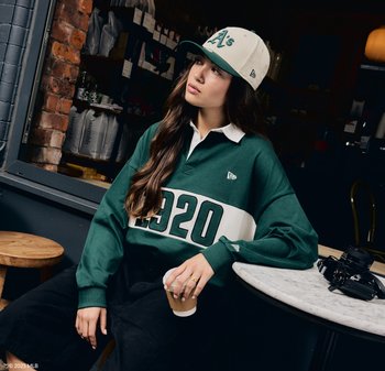 Maglietta verde in stile rugby con una striscia bianca e la stampa "1920", abbinata a un cappello crema con logo verde. Pantaloni corti scuri e una tazza da asporto visibili.