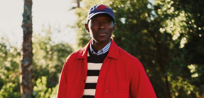 Mann, der eine rote Jacke, einen gestreiften Pullover und eine blaue Kappe trägt, steht im Freien mit Bäumen und Sonnenlicht im Hintergrund.