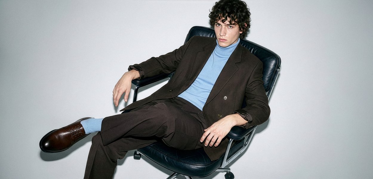 Νέος άνδρας με σγουρά μαλλιά που φοράει καφέ κοστούμι, μπλε ζιβάγκο και κάλτσες, αναπαύεται σε μαύρη γραφειακή καρέκλα με απλό λευκό φόντο.