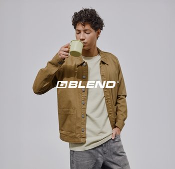 Jonge man in een bruin jasje en grijze jeans drinkt uit een lichtgroene mok tegen een effen grijze achtergrond.