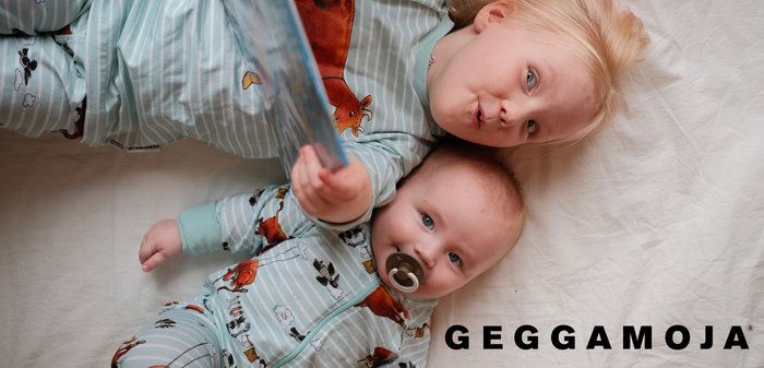 Two children lie on a light surface, wearing animal-patterned pajamas in pastel colors. One child holds a book, while the other has a pacifier.
