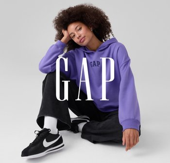 Lila kapucnis pulóver „GAP” logóval, fekete farmer és fekete Nike tornacipő fehér díszítéssel. A modell ül, göndör hajjal.