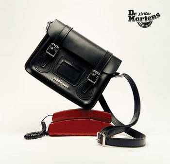 Zwarte leren crossbodytas met een klep, twee metalen gespen en een zak; heeft een gladde textuur en verstelbare riem. Rode vintage telefoon eronder.