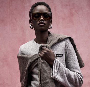 Persona che indossa occhiali da sole marroni oversize, orecchini a cerchio d'argento, maglione grigio con toppa "WRSTBHVR", che tiene una giacca marrone sulla spalla.