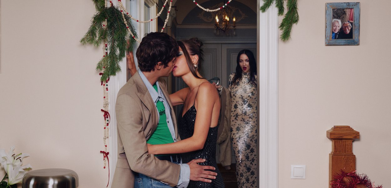 Ζευγάρι φιλιέται κάτω από γαρνιτούρα διακοπών στην πόρτα, γυναίκα με φορεσιά από παγιέτες πίσω τους με σοκαρισμένη έκφραση.