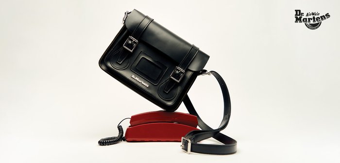 Borsa a tracolla in pelle nera con fibbie argentate lucide, forma rettangolare e logo impresso. Posata su un telefono retrò rosso.