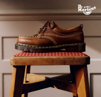 Bruine leren Dr. Martens-schoen met gele stiksels op een rode, met kussen beklede houten kruk tegen een beige, gepleisterde muur.