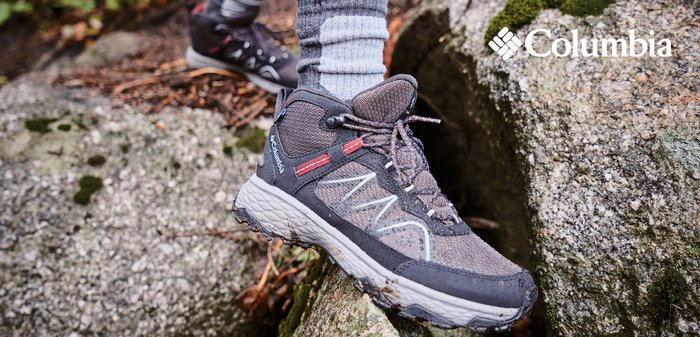 Grauer Wanderschuh mit roten Akzenten und strukturierten Oberflächen, auf felsigem Terrain platziert. Knöchelhöhe mit gepolsterter Sohle und sichtbarem Logo.