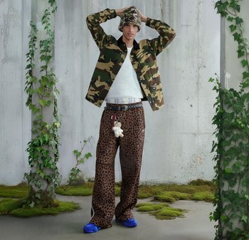 Veste camouflage, chemise blanche, pantalon à imprimé léopard avec poches, chaussures bleues et bonnet à motif, debout au milieu du feuillage vert.