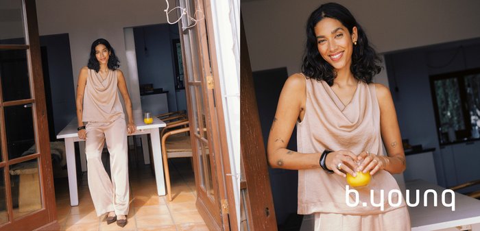 Frau in beige-farbenem Satin-Outfit, die ein Glas Orangensaft hält und sich an einen Türrahmen in einem modernen Essbereich lehnt.