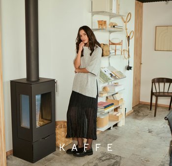 Grijze gebreide top, zwarte geplisseerde rok en zwarte schoenen; geplaatst nabij een zwarte houtkachel in een minimalistische kamer met boekenplanken en decor.