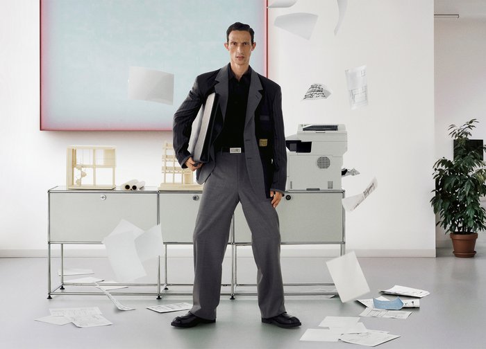 Άνδρας με επίσημη ενδυμασία στέκεται κρατώντας έναν φάκελο ανάμεσα σε διασκορπισμένα και πετώντα χαρτιά σε ένα μοντέρνο γραφείο με φωτοτυπικό μηχάνημα και φυτό.