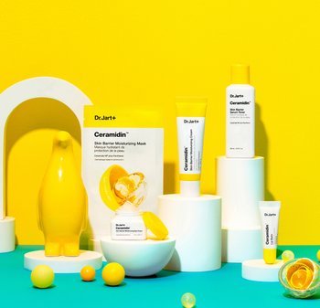Dr. Jart+ Ceramidin-produkter på vita piedestaler: en gul pingvinformad figur, en återfuktande mask, kräm och toner; ljusgul bakgrund.
