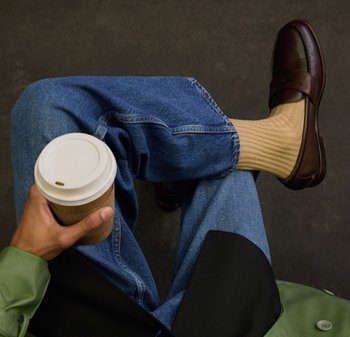 Άτομο που φοράει μπλε τζιν, μπεζ κάλτσες και σκούρα καφέ μοκασίνια, κρατώντας ένα χάρτινο ποτήρι καφέ με λευκό καπάκι, καθισμένο με σταυρωμένα πόδια.