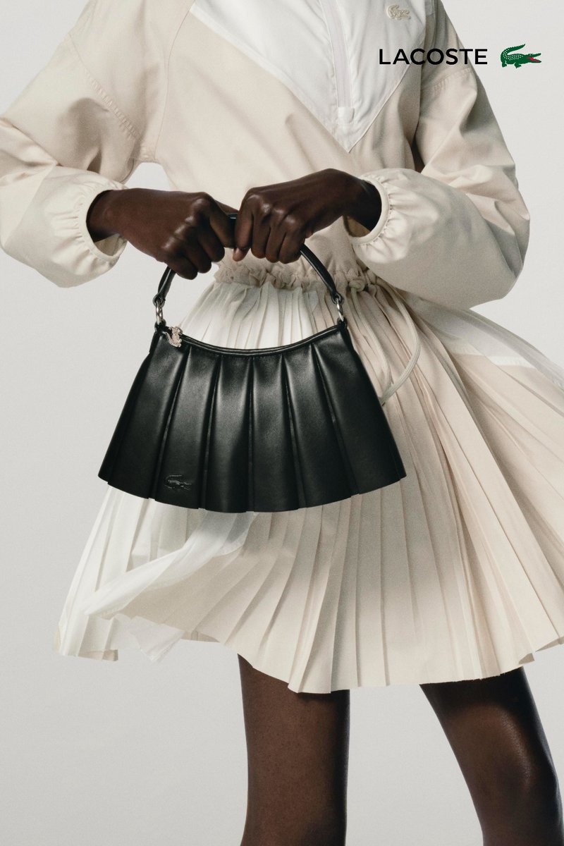 Deux mannequins posent de manière rapprochée, l'un portant un costume noir et l'autre un haut orange arborant la marque "LACOSTE", tenant des sacs élégants sur fond uni.
