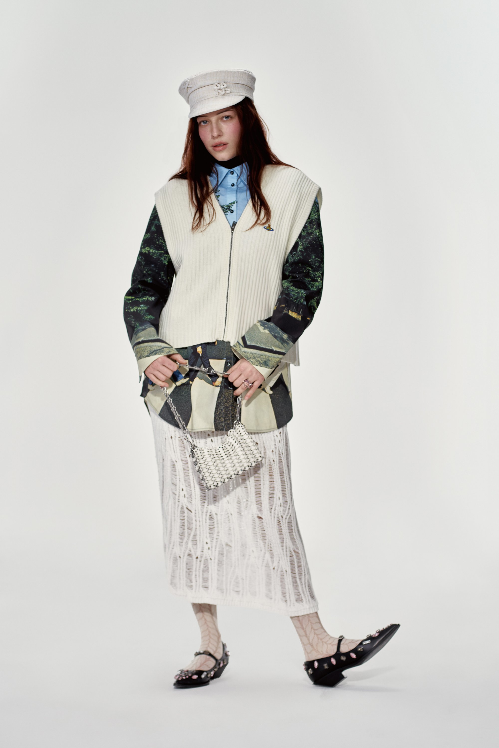 La modelo posa con un conjunto de capas que incluye un chaleco blanco, una camisa azul, una falda estampada y unos zapatos negros adornados, sosteniendo un pequeño bolso metálico.