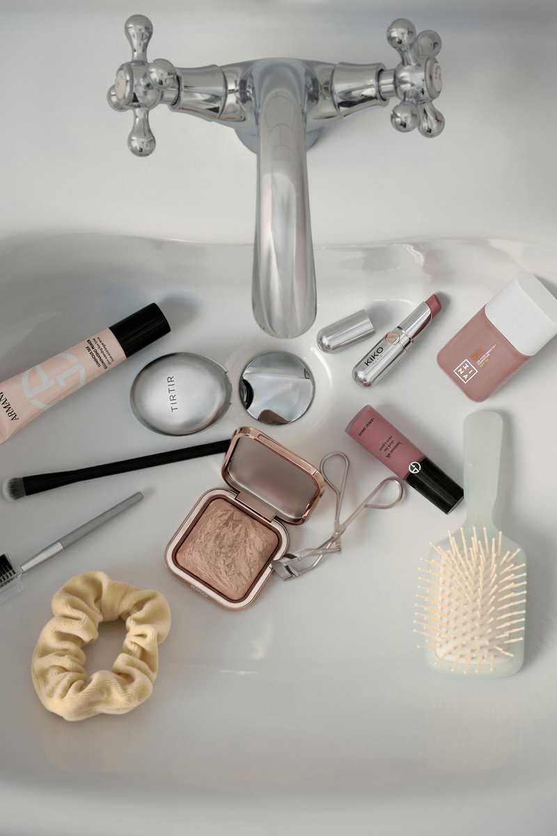 Rubinetto argentato sopra il lavandino con oggetti per il trucco sparsi, spazzola per capelli, piegaciglia e elastico per capelli all'interno della vasca.