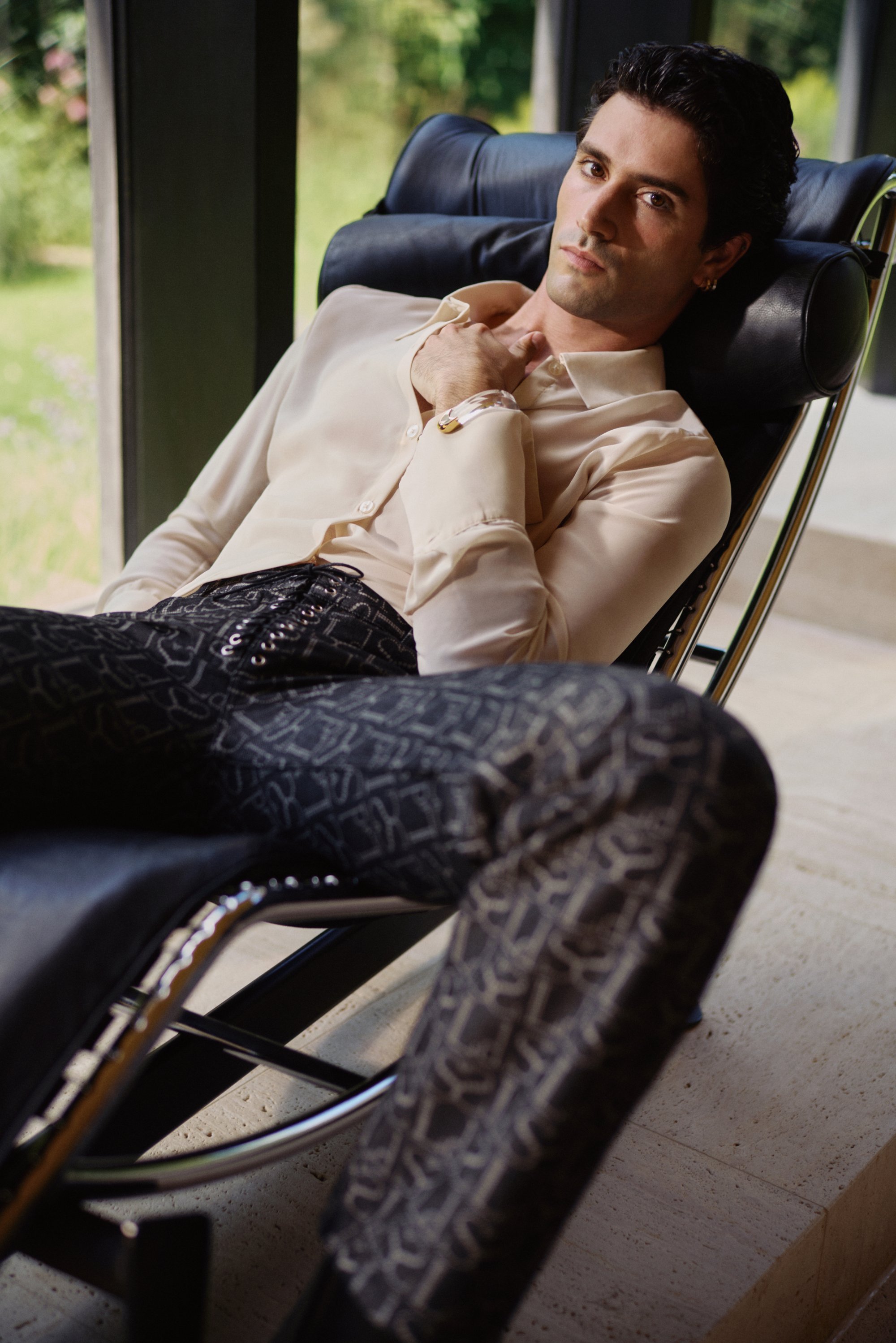 A man lounges on a sleek black chair, wearing a light button-up shirt and patterned pants, striking a contemplative pose in a bright interior.