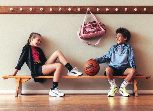 To barn sitter på en benk i en idrettsarena. Den ene har svart jakke, rød skjorte, shorts og hvite joggesko. Den andre har blå genser, shorts og sko med gult påtupp, og holder en basketball. En rosa Puma-veske henger på veggen over dem.