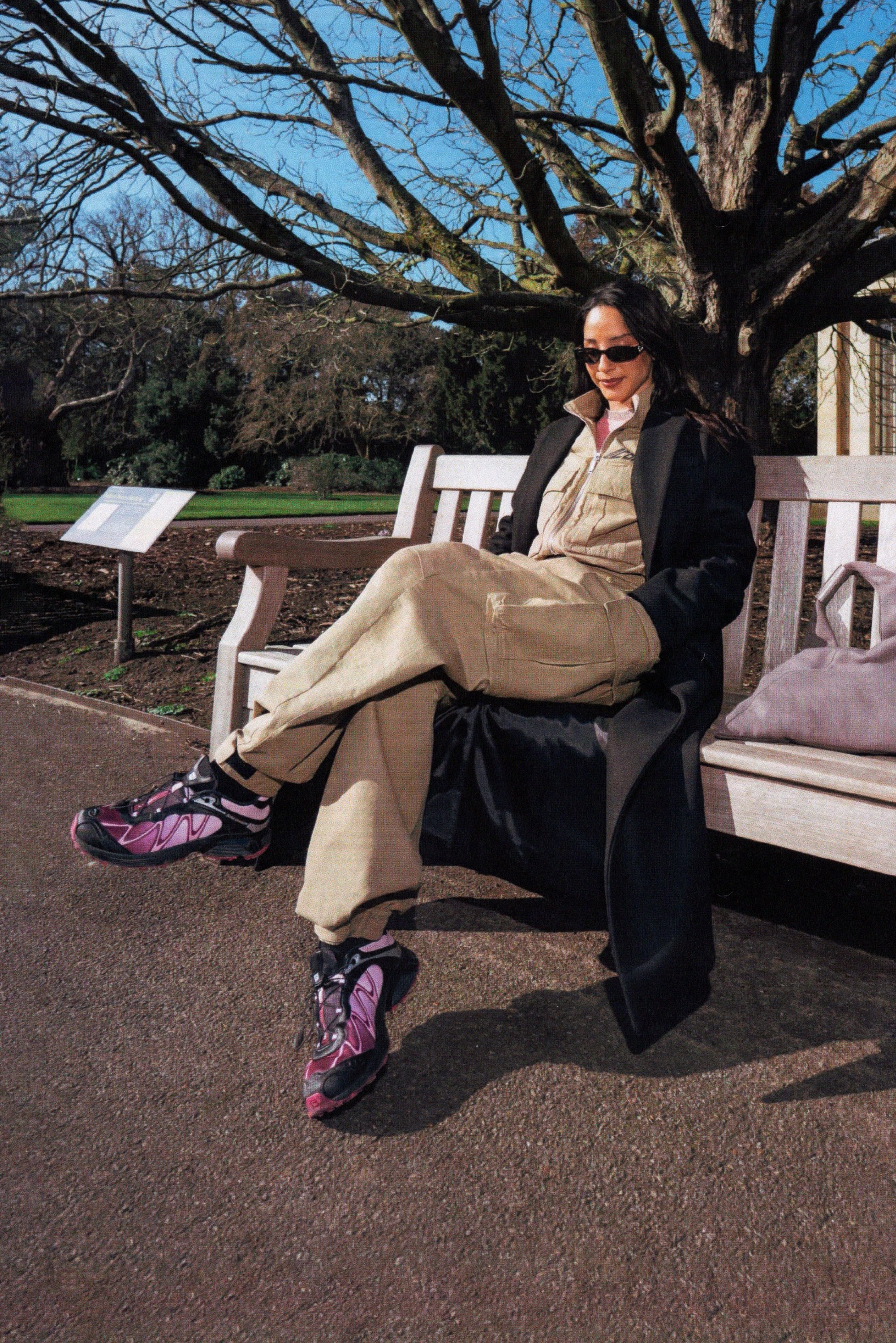 Een vrouw zit op een bankje in een park, gekleed in een beige outfit en een zwarte jas. Ze draagt stijlvolle roze sneakers, met bomen en een helder blauwe lucht op de achtergrond.
