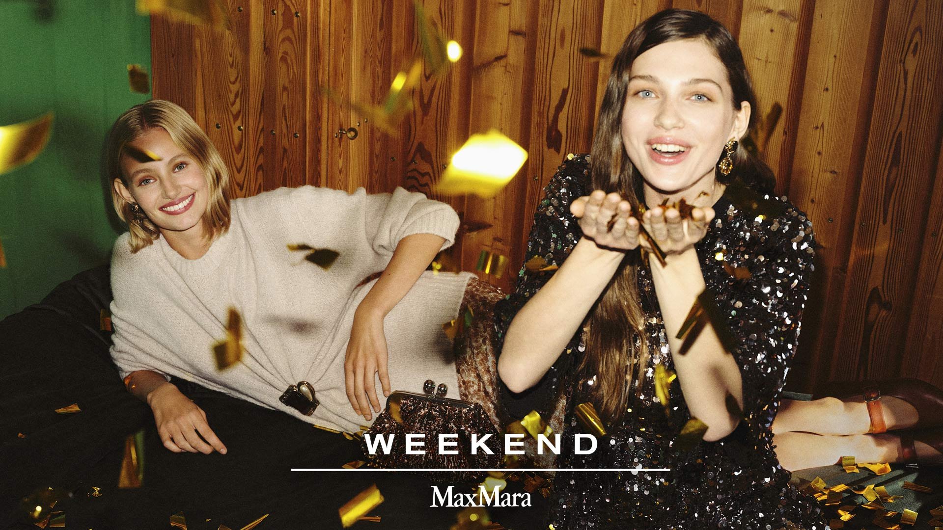 Kaksi naista istuu tummalla sohvallaan, toinen vaaleassa neuleessa ja toinen mustassa paljettimekossa. Kultaiset konfetit leijuvat ilmassa puupaneeliseinän edessä.