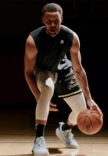 A male basketball player dribbles a ball on court, wearing a sleeveless shirt and legging, focused and in motion under dramatic lighting.