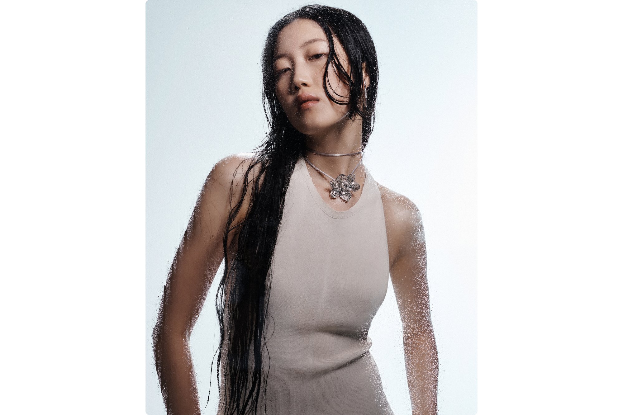 A woman with long, damp black hair poses confidently, wearing a fitted beige dress and a silver flower necklace, set against a misty background.