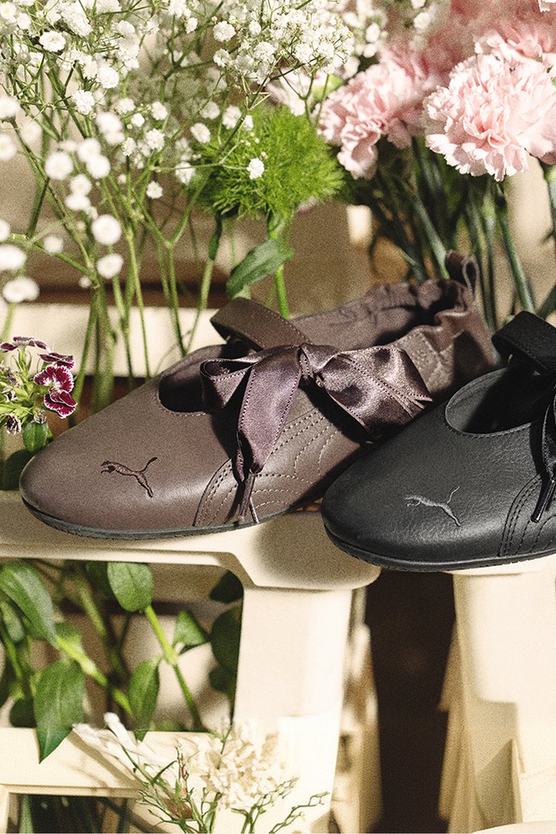 Dos bailarinas Puma en marrón y negro con lazos de cinta, exhibidas en una silla blanca rodeada de varias flores en tonos pastel.