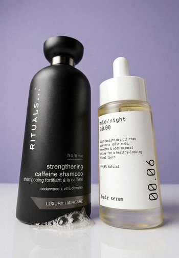 Zwart flesje met verstevigende cafeïne shampoo met schuim naast doorzichtige pipetfles met lichte haarserum op paarse achtergrond.