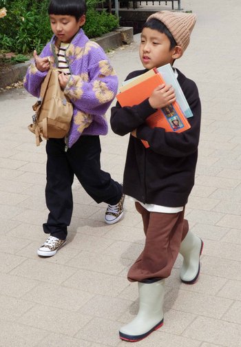 Deux enfants marchent sur un chemin pavé, l'un portant une veste fleurie violette avec un sac à dos beige, l'autre un chapeau marron tenant des livres et portant des bottes de pluie.