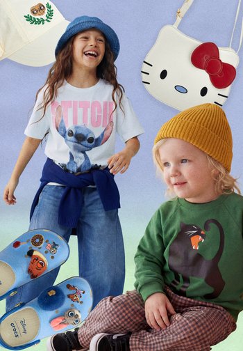 Lächendes Kind mit blauer Mütze, Stitch-T-Shirt und Jeans steht neben Kleinkind mit senffarbener Mütze, grünem Sweatshirt mit Katzenmotiv und karierter Hose.