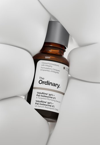 Brązowa butelka z serum z etykietą The Ordinary Volufiline 92% + Pal-Isoleucine 1%, trzymana między gładkimi, białymi, abstrakcyjnymi kształtami.