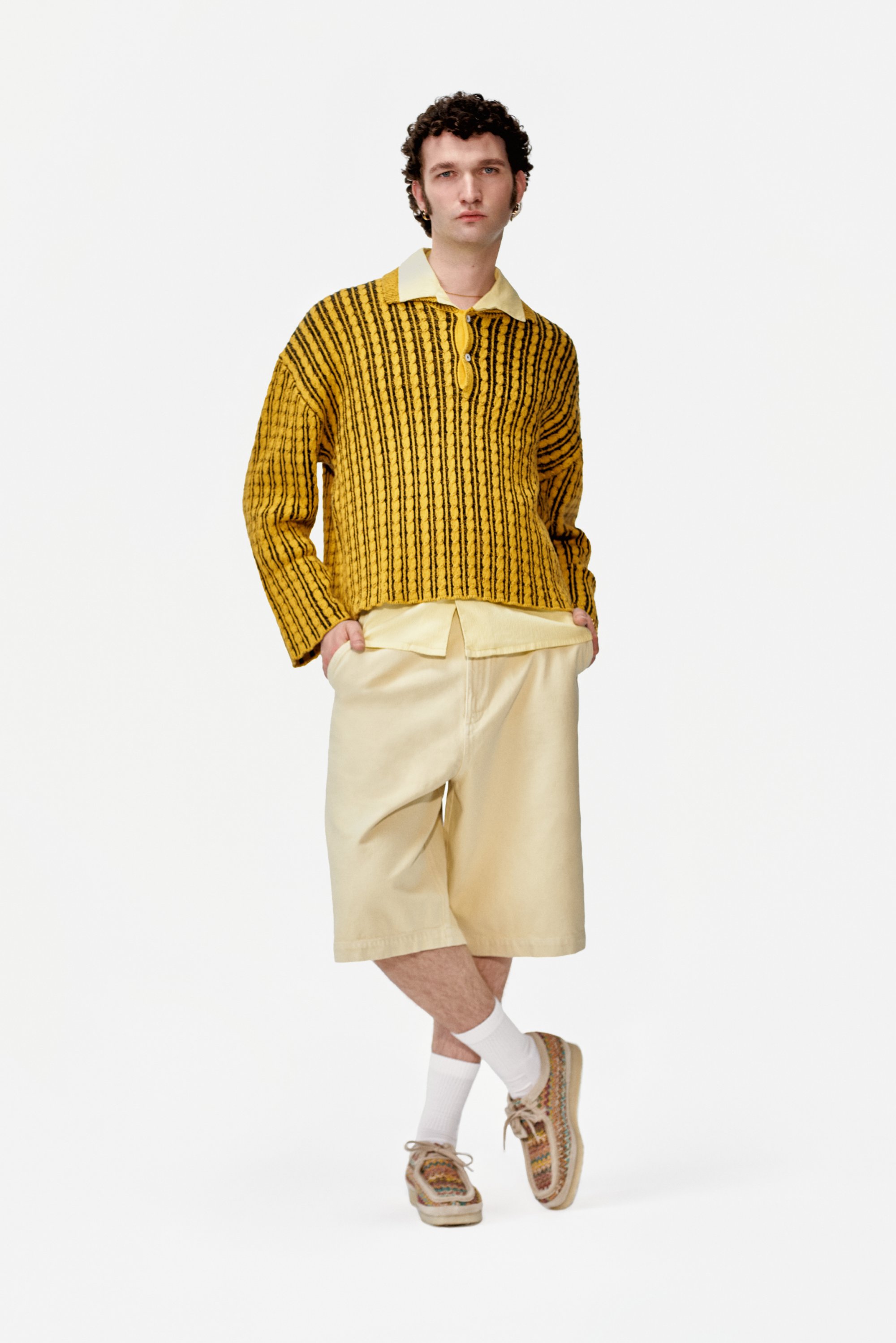 A young man stands with arms crossed, wearing a yellow striped sweater, beige shorts, and patterned shoes against a plain white background.