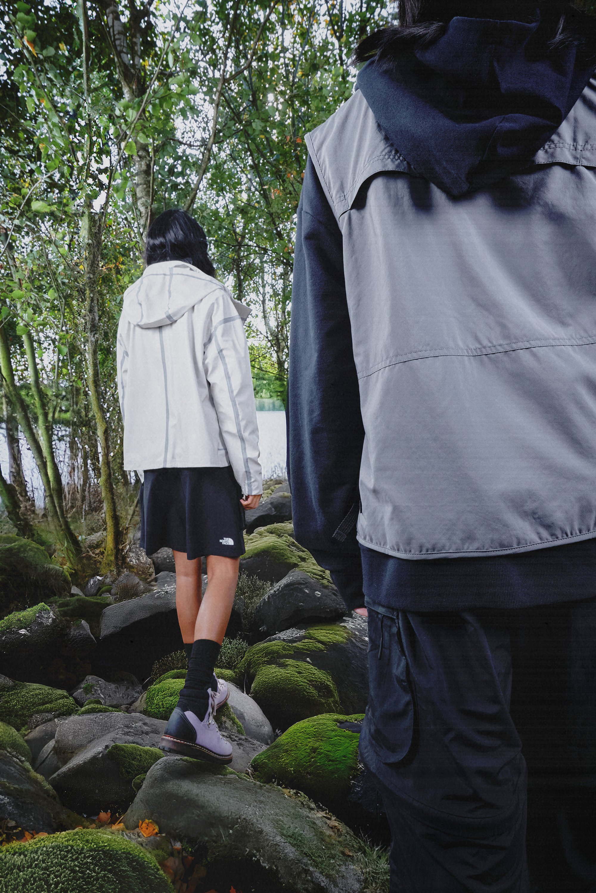 Two individuals in outdoor fashion stand on moss-covered rocks by a lake, surrounded by trees. One wears a light jacket and skirt.