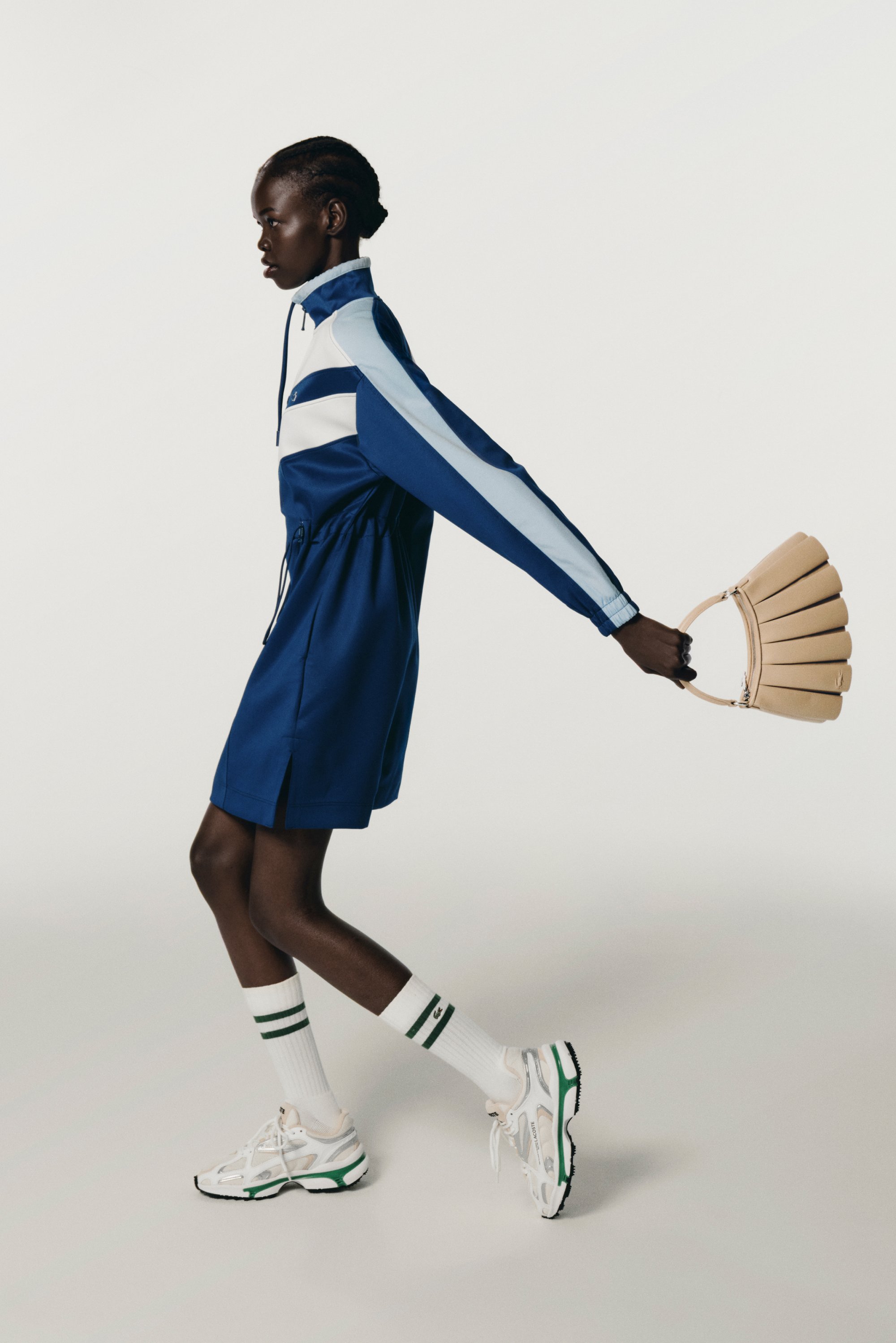 Una persona con capelli intrecciati cammina con sicurezza, indossando un completo sportivo blu e sneakers bianche, tenendo un guanto beige in una mano.
