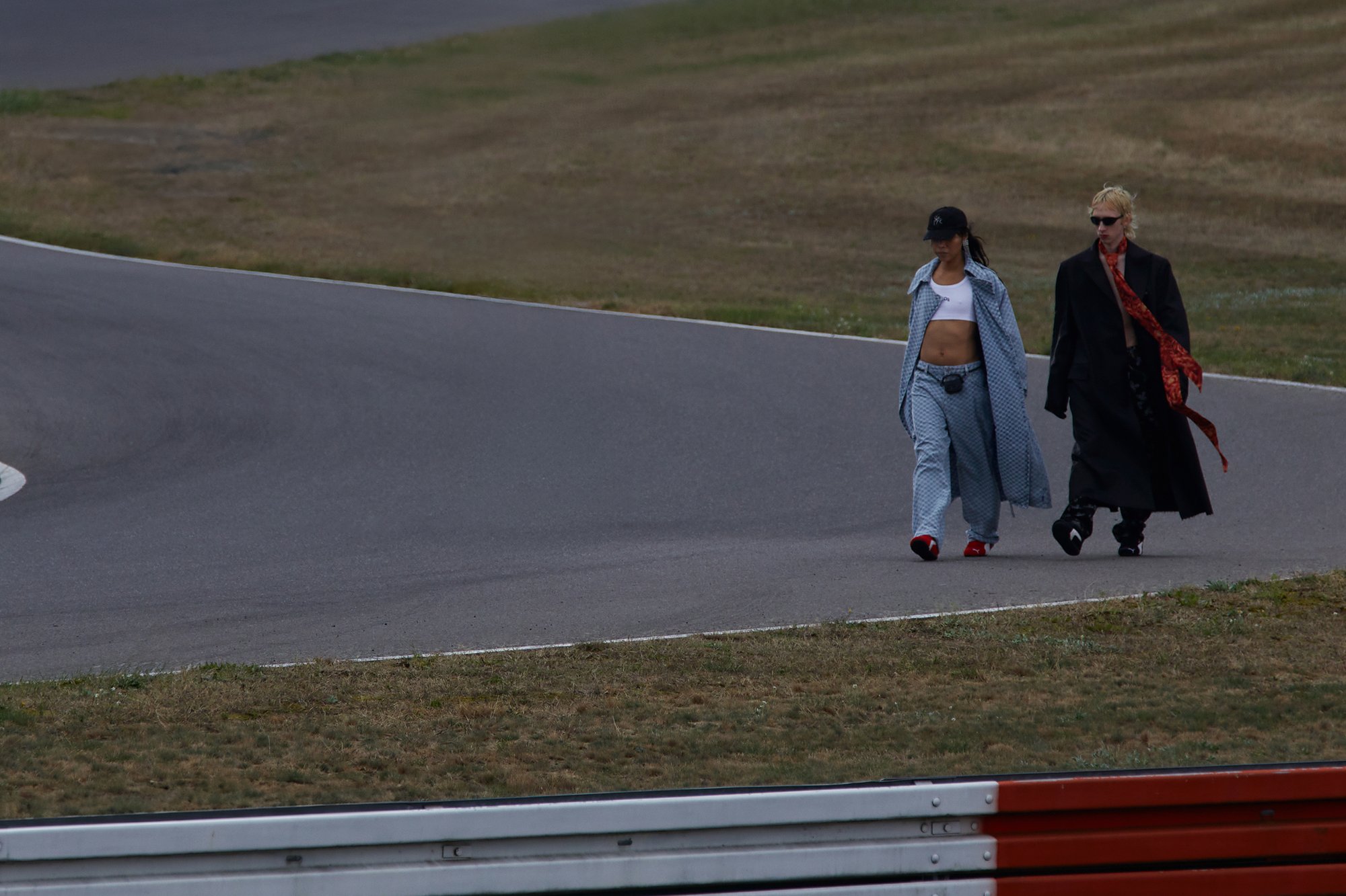 Twee personen lopen op een racecircuits, de één in een blauwe outfit en pet, de ander in een lange zwarte jas met een rode sjaal, tegen een grasrijke achtergrond.