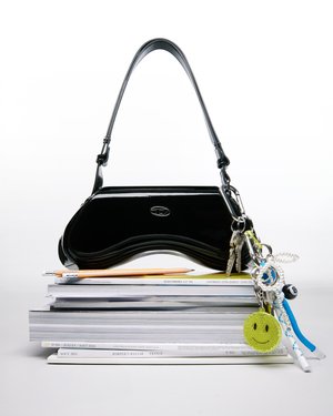 A glossy black handbag with a silver logo rests atop a stack of magazines, alongside a pencil and colorful keychains.