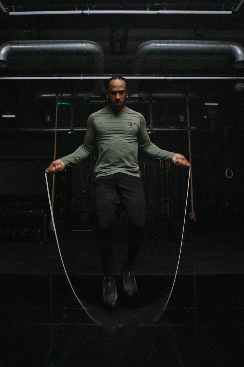 A person with braided hair wears a green long-sleeve top and black pants, jumping rope in a dark gym with fitness equipment in the background.